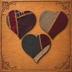 May include: Three heart-shaped fabric ornaments featuring patchwork designs in shades of burgundy, charcoal, and tan. Each heart is constructed from multiple fabric pieces joined by visible yellow cross-stitch embroidery detailing along the seams.