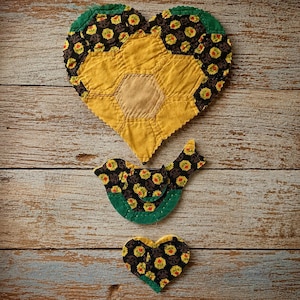 May include: Three handmade fabric items: a large heart, a bird, and a small heart. The heart and bird shapes feature a black floral pattern with yellow and red accents, and green trim. The large heart is yellow with a tan center.