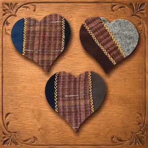 Puede incluir: Tres adornos de tela en forma de corazón con diseños de patchwork. Cada corazón combina tela de cuadros marrón con secciones lisas en azul marino, marrón oscuro o gris, unidas por bordados de punto de cruz amarillo en las costuras.