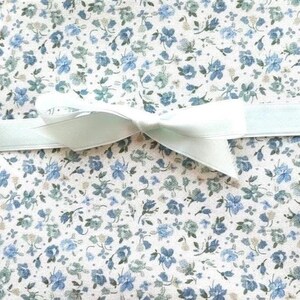 Puede incluir: Primer plano de una caja de regalo con un estampado floral en tonos azules y verdes sobre un fondo blanco. Una cinta azul claro está atada en un lazo en la parte superior de la caja, añadiendo un toque decorativo.