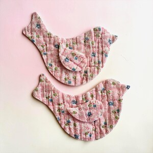 May include: Two pink fabric bird-shaped decorations with a floral pattern in blue, green, and white. The birds are quilted and have a small wing detail. The background is a gradient of pink and white.