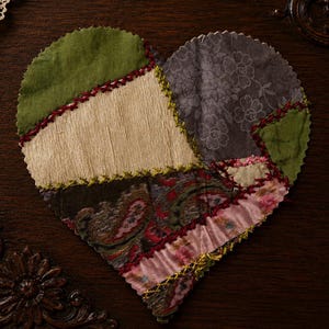 May include: A heart-shaped patchwork textile with various fabric patterns and colors. The design includes green, beige, gray floral, and paisley fabrics, stitched together with red and yellow thread. The item is likely a decorative piece or coaster.