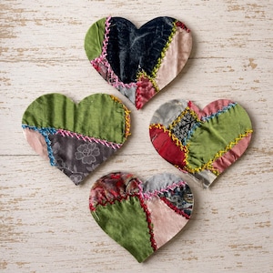 May include: Four heart-shaped fabric ornaments featuring a patchwork design. Each heart is constructed from various textile scraps in shades of green, pink, black, and red, joined together with visible, colorful cross-stitch embroidery thread.