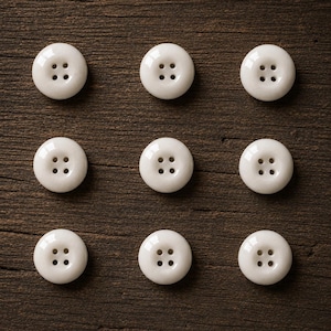 May include: Nine round, white buttons arranged in a three-by-three grid. Each button features a smooth, glossy surface and four small holes positioned in the center for attachment. The buttons are uniform in size and shape, displaying a clean design.