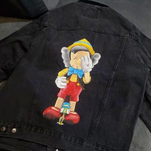 May include: Black denim jacket featuring a colorful cartoon illustration of Pinocchio with a unique artistic style. The artwork includes a yellow hat, blue bow tie, red pants, and white gloves. The jacket has a classic button-up design.