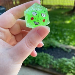 May include: A translucent neon green twenty-sided die featuring a hand-painted illustration of a face with dark outlined eyes, eyebrows, a nose, a mouth, and two red circular cheeks on one of its triangular faces, with numbers embossed on the surface.