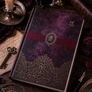 Puede incluir: Un libro de tapa dura con una cubierta texturizada de color morado oscuro y una banda horizontal de color rojo oscuro en el centro. Un emblema ovalado dorado con las letras MT se sitúa sobre la banda. El texto MAISON TETE aparece en la esquina.