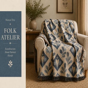 Puede incluir: Una manta tejida con un patrón de diamantes repetitivo con motivos florales azules y beige, terminada con bordes de flecos. La manta está colocada sobre un sillón tapizado. El texto a la izquierda dice Maison Tête FOLK ATELIER.