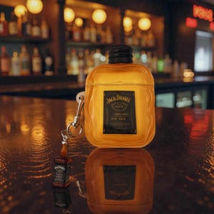 Puede incluir: Una funda transparente de color ámbar con forma de botella de whisky, con la etiqueta "Jack Daniel's". Un dije de botella en miniatura cuelga de un cierre plateado. La funda está sobre una superficie reflectante.