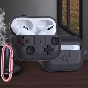 May include: Black protective case for wireless earbuds, designed to resemble a video game controller. The case features a D-pad, buttons, and a green indicator light. A rose gold carabiner clip is attached for portability. The earbuds are white.