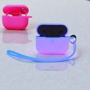 Könnte beinhalten: Zwei Schutzhüllen aus Silikon für kabellose Ohrhörer. Eine ist hellrosa und offen, die andere ist lila und offen, mit einer blauen Handschlaufe. Die Hüllen schützen die Ohrhörer vor Kratzern und Beschädigungen.