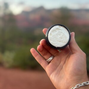 Peut inclure: Un petit récipient rond noir rempli de crème blanche. Le récipient est tenu dans une main ouverte, avec un arrière-plan naturel flou de montagnes et de ciel. Une bague et un bracelet en argent sont également visibles.