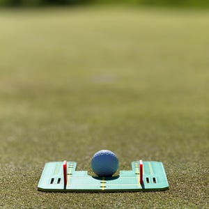 May include: A golf ball sits on a green putting alignment tool with red and yellow markings. The tool is on a green grassy surface. The golf ball is light blue and has dimples.