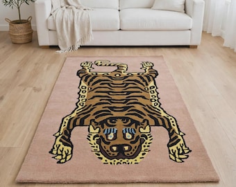 May include: A rectangular area rug featuring a stylised tiger skin design in shades of brown, black, and yellow on a solid dusty pink background. The tiger motif is centred, displaying a flat, symmetrical pattern with distinct stripes and paws.