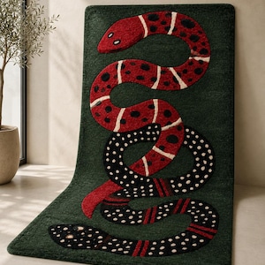 May include: A rectangular dark green rug featuring a large, coiled snake design. The snake has a red upper body with black spots and white bands, transitioning into a black lower body with white spots and red bands, all set against a solid green background.