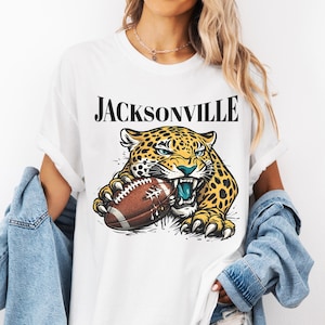 May include: A white t-shirt featuring the text JACKSONVILLE in black serif font above a graphic illustration of a yellow and black spotted jaguar biting into a brown football with white laces. The jaguar has blue eyes and teeth.