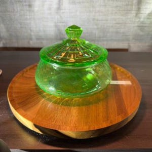 May include: A translucent, vibrant green glass candy dish with a matching lid. The bowl features a rounded base with subtle vertical ribbing, while the lid has a tiered, circular design with a small, pointed finial at the very center.