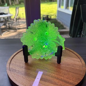 May include: A vibrant, translucent neon green glass dish shaped like a detailed grape leaf with prominent veins and three small raised circular accents. The dish is held upright by a black plastic display stand on a round wooden base.
