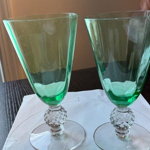 May include: Two green-tinted wine glasses. The bowls are a trapezoidal shape, and the stems feature a decorative, textured design. The bases are round and clear. The glasses are set on a white surface.