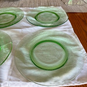 May include: Four green glass plates with a floral pattern are arranged on a white cloth. The plates have a raised, circular center and a decorative edge. The plates are on a wooden table.