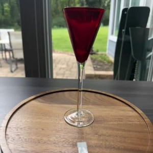 May include: A tall, slender glass with a deep red, conical bowl and a clear, thin stem resting on a circular base. The glass is positioned upright on a round, light brown wooden surface with a visible grain pattern and a small white label.