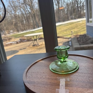 May include: A green glass candlestick holder with a round base and a flared top, sitting on a round wooden tray. The candlestick holder is made of transparent green glass. The tray is made of wood.