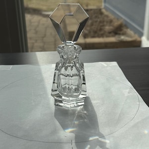 May include: A clear glass perfume bottle with a faceted, diamond-shaped stopper. The bottle has a square base and a detailed, ornate design. The bottle is sitting on a white surface, casting a shadow.