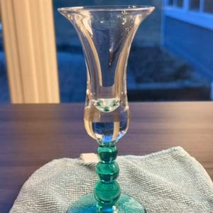 May include: A clear glass vase with a flared top and a teal green stem. The stem features a series of rounded beads. The vase is sitting on a light blue cloth.
