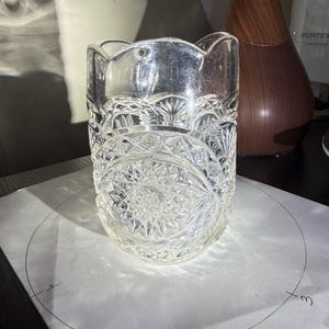 May include: A clear, cut-glass vase with a scalloped rim. The vase features a detailed, geometric pattern around the body. The vase is sitting on a white surface with a drawn circle.