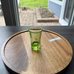 May include: A small, green shot glass sits on a round, wooden serving tray. The tray has a dark brown finish and a raised edge. The shot glass is empty and appears to be made of glass. The background is a blurred view of a backyard.