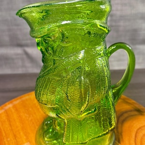 May include: A vibrant lime green glass pitcher featuring a molded, textured design of overlapping feathers or leaves. The pitcher has a curved handle, a wide pouring spout, and a circular base, crafted from translucent glass with a glossy finish.