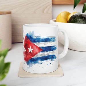 Puede incluir: Taza de cerámica blanca con un diseño de acuarela de la bandera cubana. El diseño de la bandera incluye un triángulo rojo con una estrella blanca, rayas azules y blancas y salpicaduras de acuarela. La taza está sobre un posavasos beige.