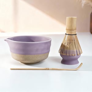 May include: A matcha tea set comprising a lavender ceramic bowl with a beige base, a bamboo whisk, and a lavender whisk holder. The set is designed for preparing matcha tea. The items are arranged on a white surface.