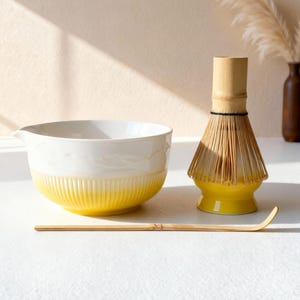 以下が含まれることがあります： 白と黄色のセラミックボウル、竹の泡立て器、竹のスクープを備えた抹茶ティーセット。 ボウルはリブ付きの黄色のベースと白い上部セクションがあります。 抹茶の準備用に設計されたセットです。