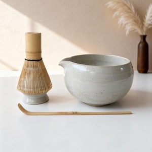 May include: A matcha tea set featuring a ceramic bowl with a spout, a bamboo whisk, and a bamboo scoop. The bowl and whisk base are light gray with speckled details. The whisk is made of thin bamboo tines.