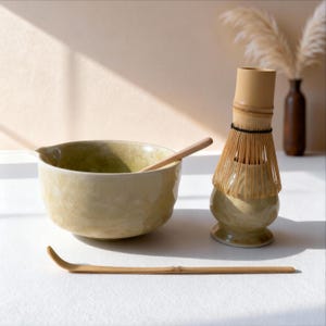 May include: A matcha tea set featuring a ceramic bowl with a wooden spoon, a bamboo whisk, and a bamboo scoop. The bowl has a light green interior and a beige exterior. The whisk and scoop are made of bamboo. The set is on a white surface.