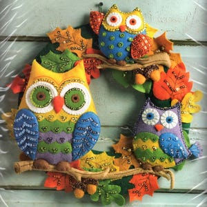 May include: A decorative wreath featuring three felt owls in yellow, blue, and purple, adorned with sequins. The owls are perched on felt branches surrounded by orange, yellow, and green felt autumn leaves and small brown felt acorns.
