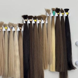 May include: A collection of hair extensions in various shades, from light blonde to dark brown and black, are displayed. Each bundle is secured with a small band and hung on a white rack against a white wall. The hair appears smooth and straight.