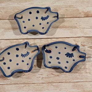 May include: Three ceramic pig-shaped dishes. Each dish is light beige with blue polka dots and a blue rim. The pig's features, including closed eyes and snout, are also in blue. The dishes are on a light wood surface.