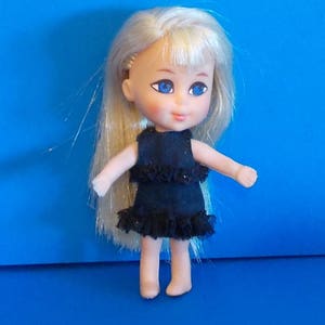 May include: A small doll with blonde hair and blue eyes, wearing a black dress with ruffled trim. The doll is standing against a solid blue background. The doll's arms are outstretched.