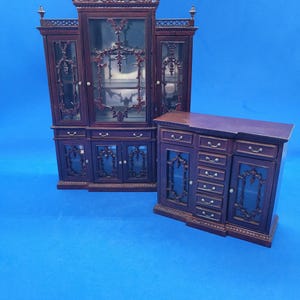 May include: Two miniature wooden cabinets with a dark brown finish. One is a tall, ornate display cabinet with glass doors and decorative carvings. The other is a shorter cabinet with drawers and glass-fronted doors. Both are on a blue background.