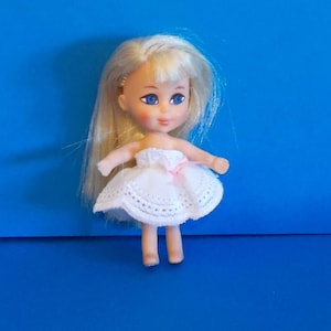 May include: A small, white doll with blonde hair and blue eyes. She is wearing a white dress with lace trim and a pink ribbon.