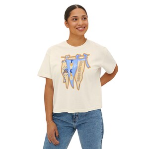 May include: Cream-colored, short-sleeved t-shirt with a graphic of three pennant flags. The flags are light blue and tan, with the words "TRUE GOLDEN HARE" printed on them. The shirt is paired with blue jeans.