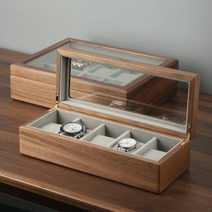 May include: A wooden watch box with a clear glass top, showcasing two watches. The box is open, revealing gray padded compartments. The wood has a warm, natural grain. Another closed box is in the background.
