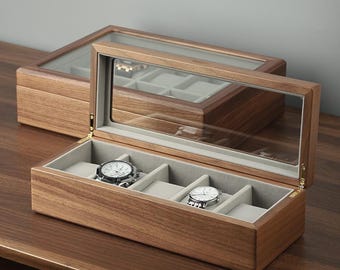 May include: A wooden watch box with a clear glass top, showcasing two watches. The box is open, revealing gray padded compartments. The wood has a warm, natural grain. Another closed box is in the background.