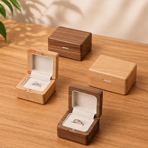 May include: Four wooden ring boxes, two open to reveal rings. The boxes are rectangular with a small white accent on the front. Two boxes are light wood, and two are dark wood. The rings are silver and set in a white interior.