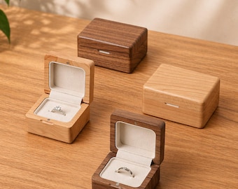 May include: Four wooden ring boxes, two open to reveal rings. The boxes are rectangular with a small white accent on the front. Two boxes are light wood, and two are dark wood. The rings are silver and set in a white interior.