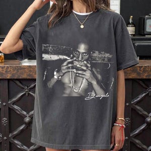 Puede incluir: Camiseta gris oscuro con una imagen en blanco y negro de una persona sosteniendo un cigarrillo y usando collares. El nombre "D'angelo" está escrito en cursiva. La camiseta tiene un corte holgado.