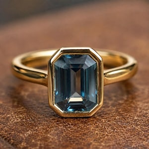 May include: A gold ring featuring a rectangular emerald-cut blue gemstone set in a smooth bezel setting. The band is polished gold with a rounded profile, holding the central stone securely in a clean, minimalist metal frame design.
