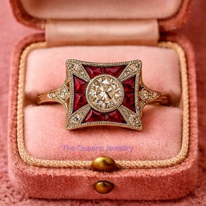 Könnte beinhalten: Ein goldener Ring mit einem quadratischen, rot und diamantbesetzten Mittelstück. Die Mitte zeigt einen großen, runden Diamanten, umgeben von kleineren Diamanten und roten Steinen. Der Ring befindet sich in einer rosa Samtbox. Text: "The Queens Jewellery."